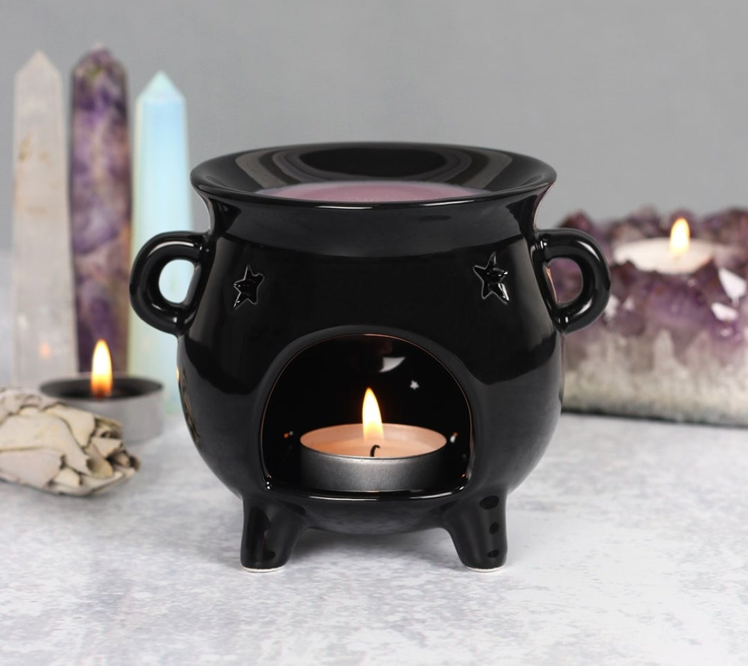 A black oil burner and wax warmer with a cut out pentagram design. It is shaped like a cauldron with a small candle lit inside it, placed on a table with candles and crystals in the background.