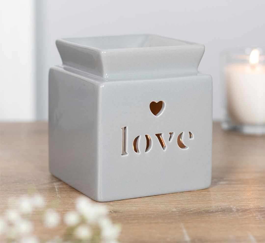A white ceramic square burner with the word 'love' cut out and a heart shape above the cutout. It is placed on a wooden surface with a lit candle and flowers in the background.
